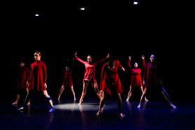 Active group of dancers in red flowing dresses mid-movement on a dark stage